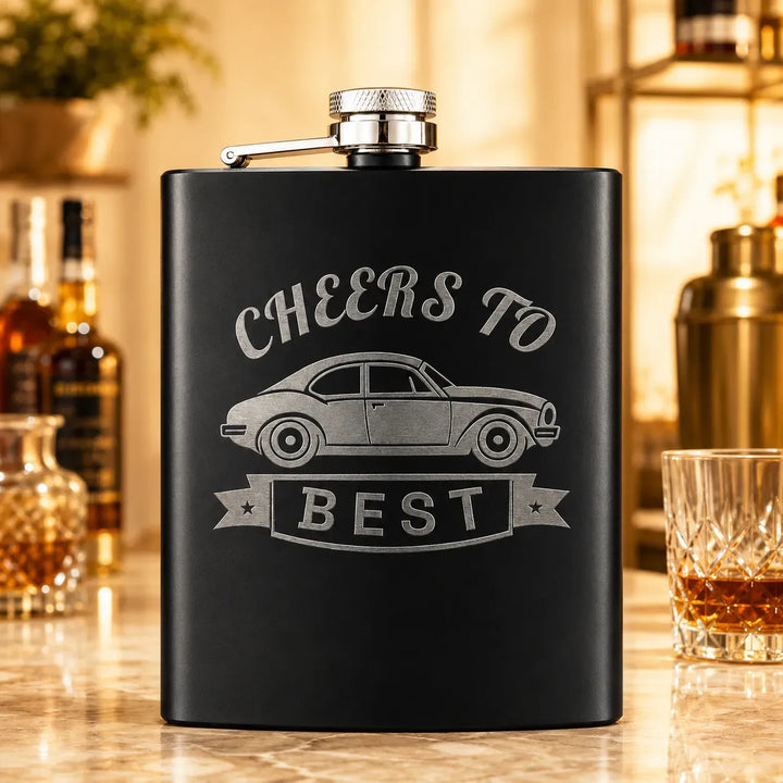 Matte black stainless hip flask laser engraved with a Cheers To The Best design in bright silver steel, on a bar top