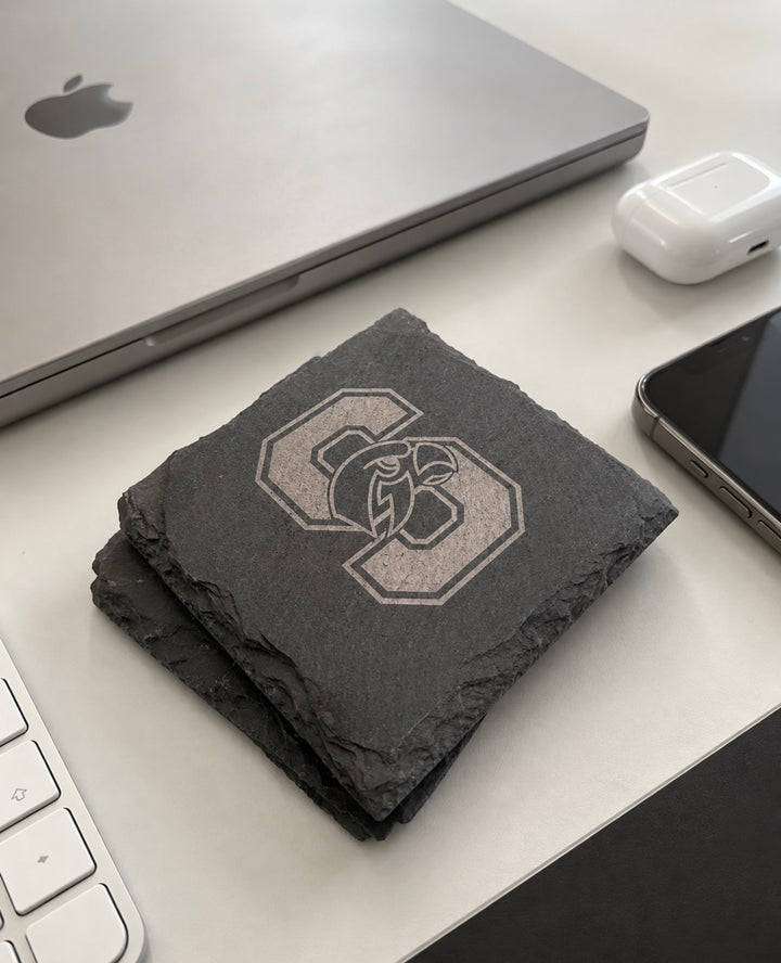 Seminole Ridge Hawks slate coasters stacked on a desk beside a laptop, phone and earbuds